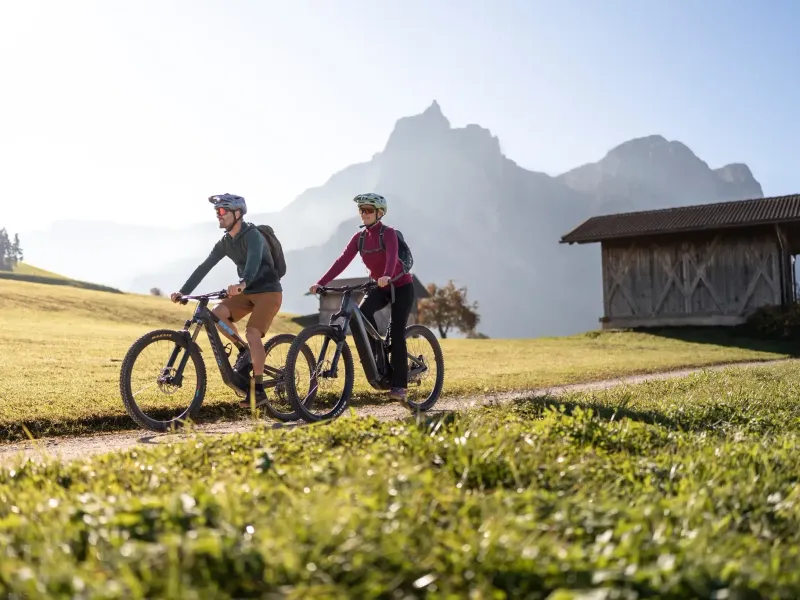 Zwei Radfahrer mit Helmen fahren auf einem Feldweg vor einer Bergkulisse und einer Holzhütte