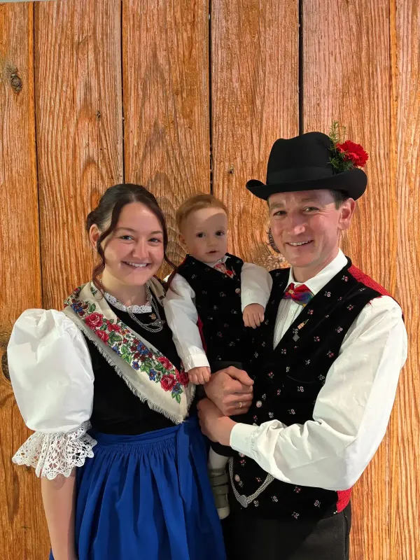 Drei Personen in traditioneller Tracht vor einer Holzpaneelwand, Frau mit weißer Bluse, besticktem schwarzem Mieder und blauem Rock, Mann mit schwarzem Hut und Weste, Kind in schwarzer Weste und weißem Hemd