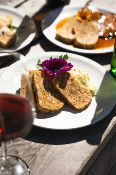 Zwei Scheiben Serviettenknödel mit Blüte und Beilage auf weißem Teller auf Holztisch mit Glas Rotwein im Vordergrund