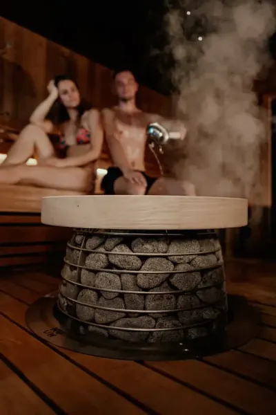 Zwei Personen in Badebekleidung sitzen in einer Sauna hinter einem Saunaofen mit aufsteigendem Dampf